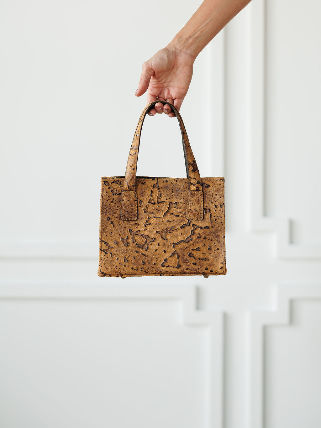 A hand holds The Nubuck, a small brown leather handbag with two handles by Savannah Sevens western life{&}style, against a white decorative wall background.