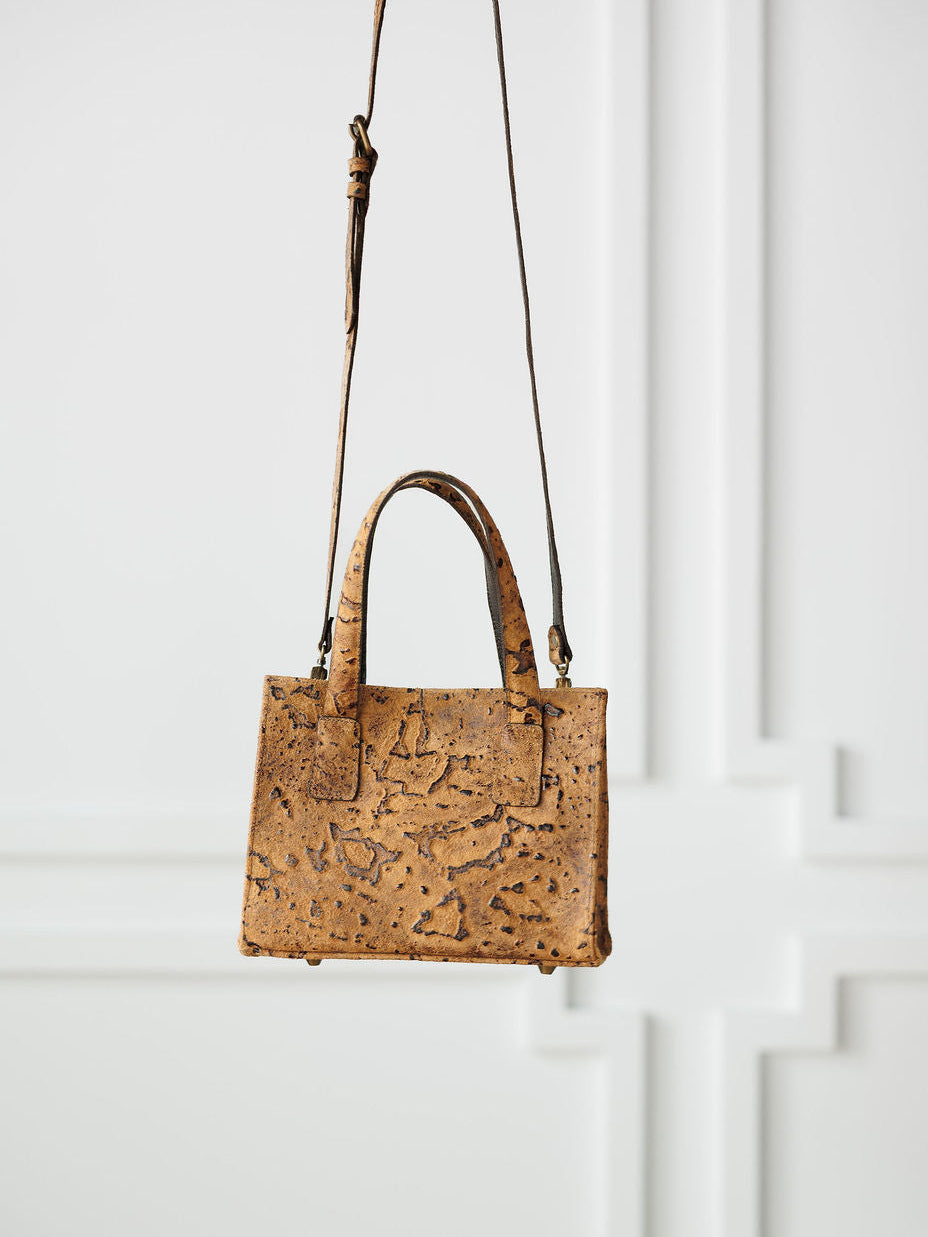 The Nubuck by Savannah Sevens western life{&}style is a small, square handmade crossbody bag with a cork texture, two handles, and a long adjustable strap, shown against a white paneled wall.