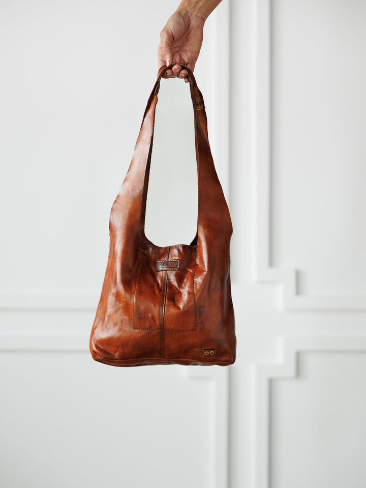 A hand holds up The Pantego by Savannah Sevens western life{&}style, a cognac leather bag with a wide strap, in front of a white wall with decorative molding.