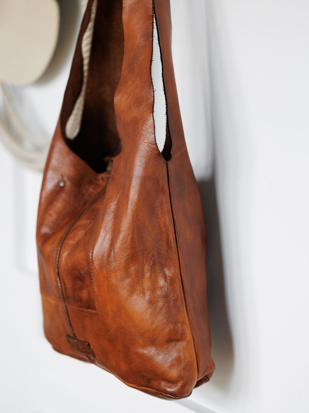 The Pantego by Savannah Sevens western life{&}style is a cognac genuine leather tote with a simple, unstructured design, featuring a smooth, slightly worn texture and a single strap, hanging on a white wall.