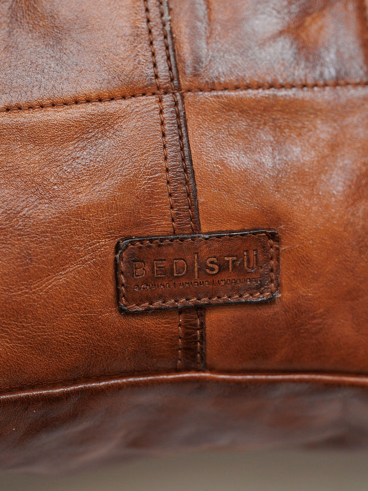 Close-up of The Pantego cognac leather bag featuring visible stitching and a rectangular patch embossed with Savannah Sevens western life{&}style and the words Genuine | Natural | Handmade.