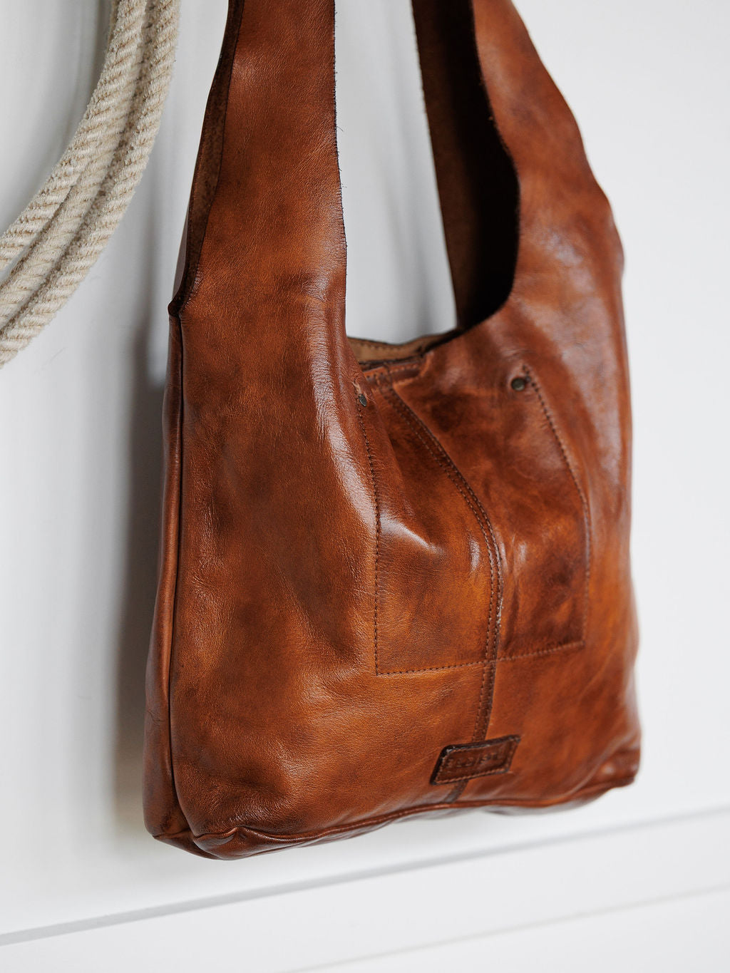The Pantego by Savannah Sevens western life{&}style, a cognac leather bag with visible stitching, hangs on a white wall beside a coiled rope.