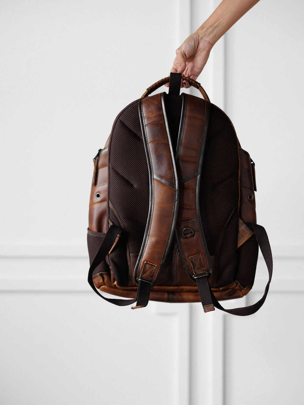 A hand holds up the Waynesboro Backpack by Savannah Sevens western life{&}style, displaying its padded back and shoulder straps against a white background.