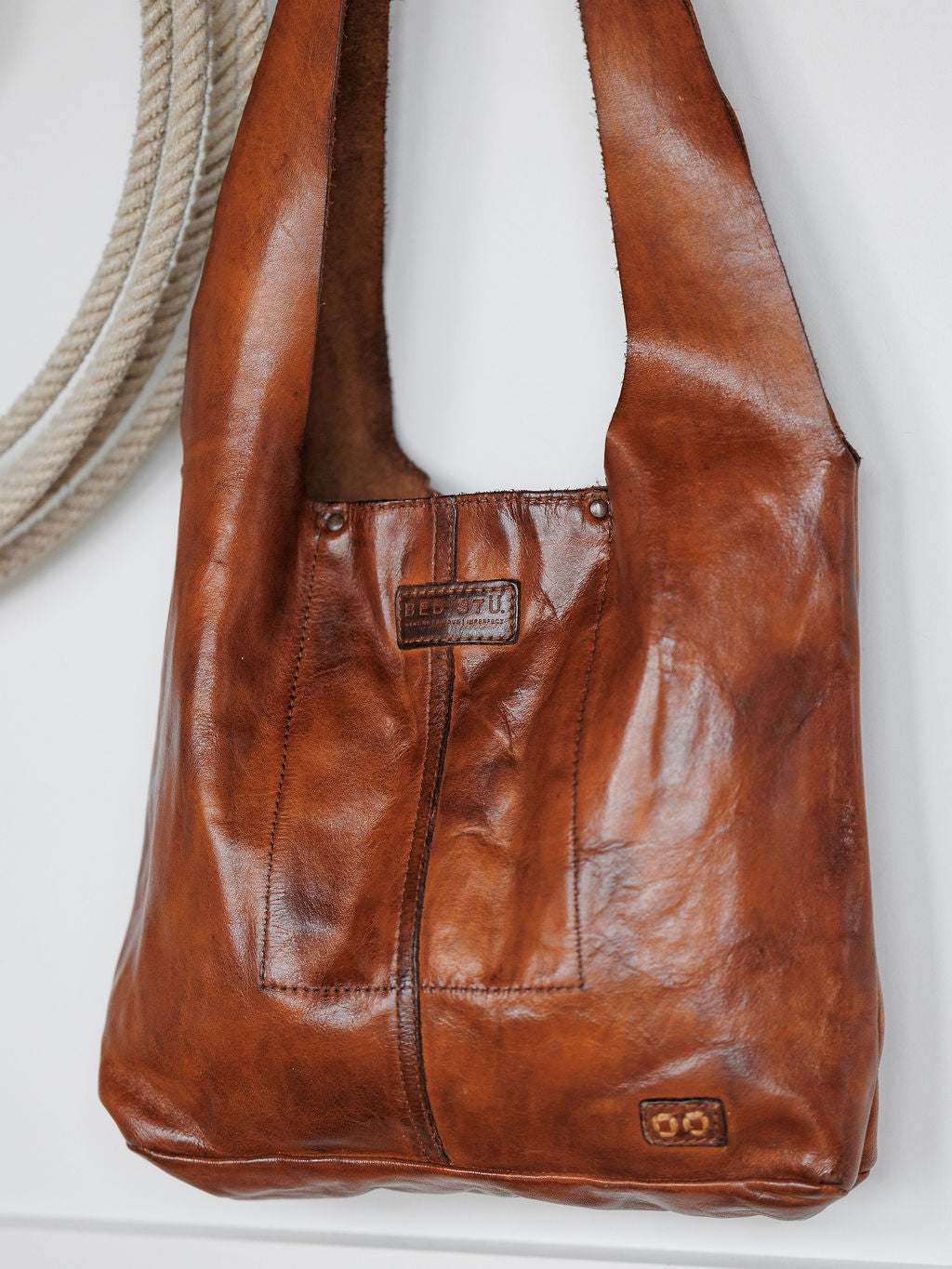 The Pantego by Savannah Sevens western life{&}style, a cognac leather tote with a natural glossy finish and visible stitching, hangs on a white wall beside a thick coiled rope. A small rectangular label accents the front.