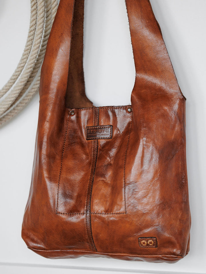 The Pantego by Savannah Sevens western life{&}style, a cognac leather tote with a natural glossy finish and visible stitching, hangs on a white wall beside a thick coiled rope. A small rectangular label accents the front.