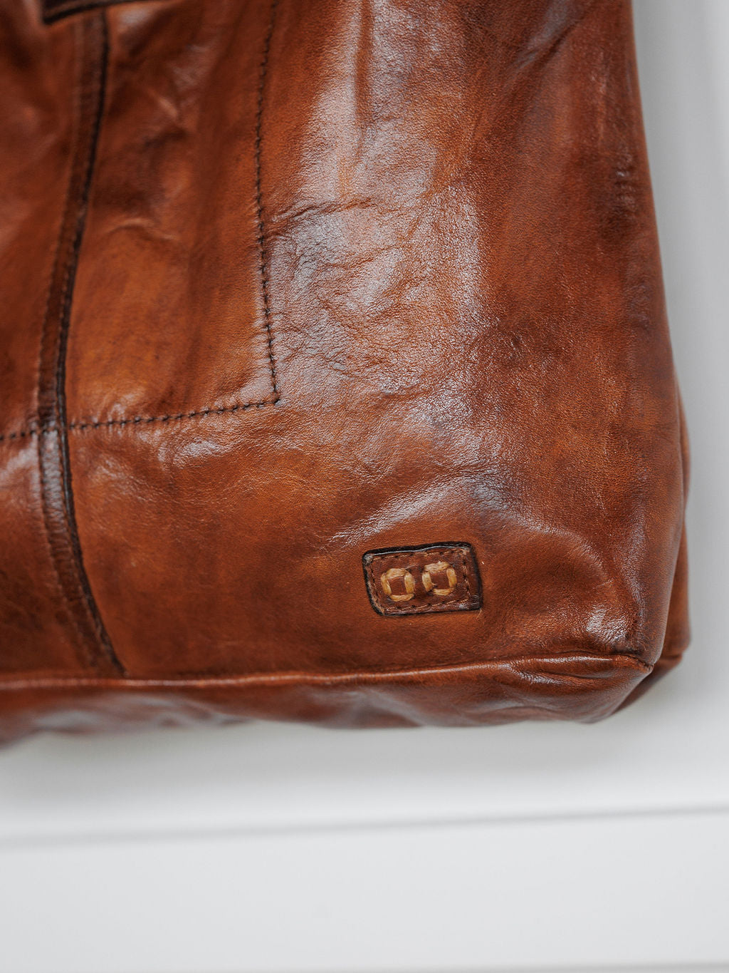 A close-up of The Pantego by Savannah Sevens western life{&}style shows its cognac leather, detailed stitching, and a small rectangular logo patch near the bottom corner, all set against a white background.