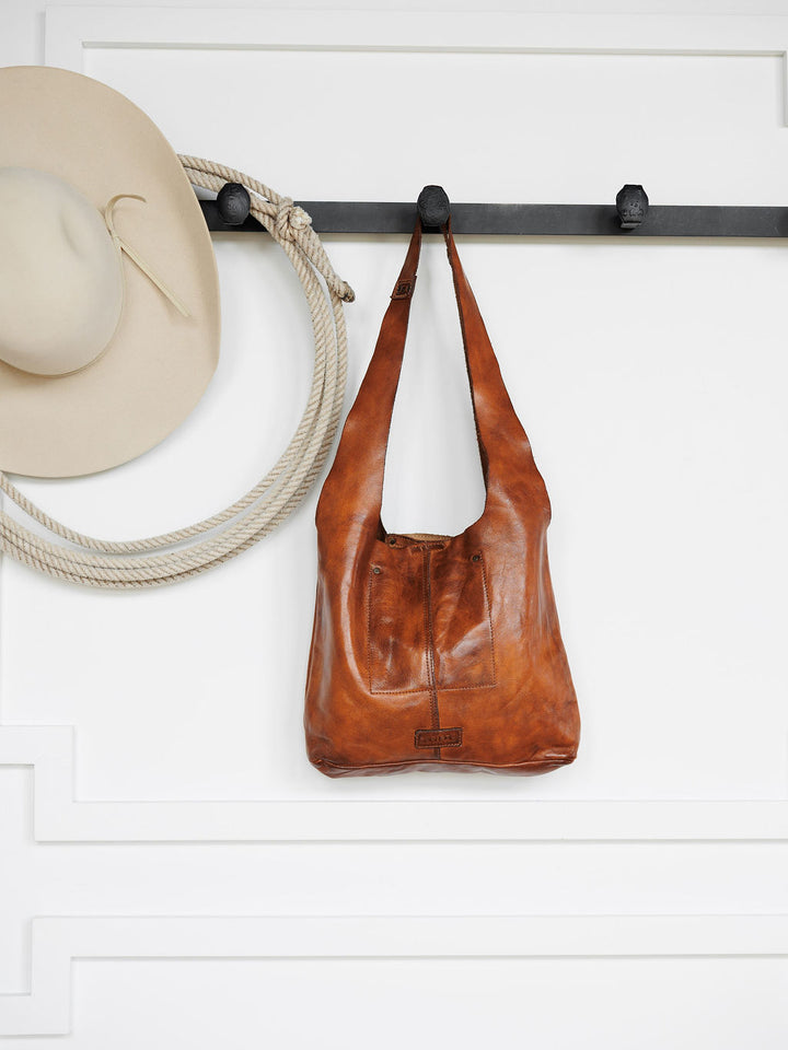 The Pantego by Savannah Sevens western life{&}style hangs from a black wall hook beside a cream cowboy hat and coiled rope, all displayed against a white wall with decorative molding.