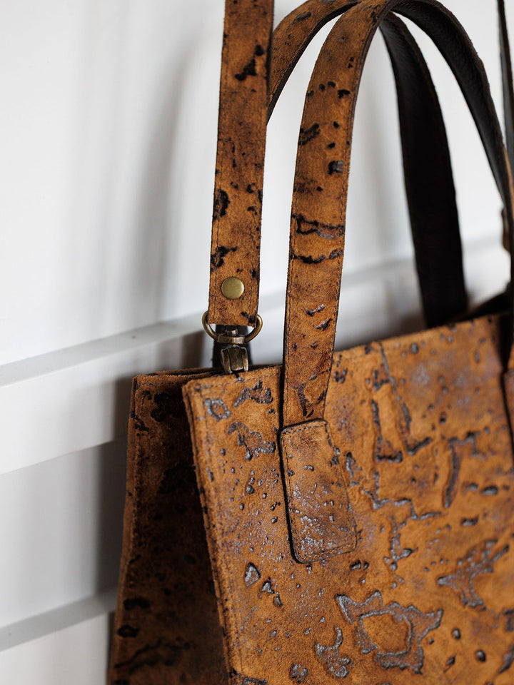 Close-up of The Nubuck by Savannah Sevens western life{&}style—a brown Nubuck leather handbag with two handles, metallic clasp, and unique mottled pattern—displayed on a white slatted wall.