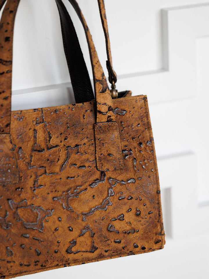 A close-up of The Nubuck by Savannah Sevens western life{&}style, a handmade nubuck leather crossbody bag with dark patterns, two handles, and a rectangular shape, shown against a white paneled background.