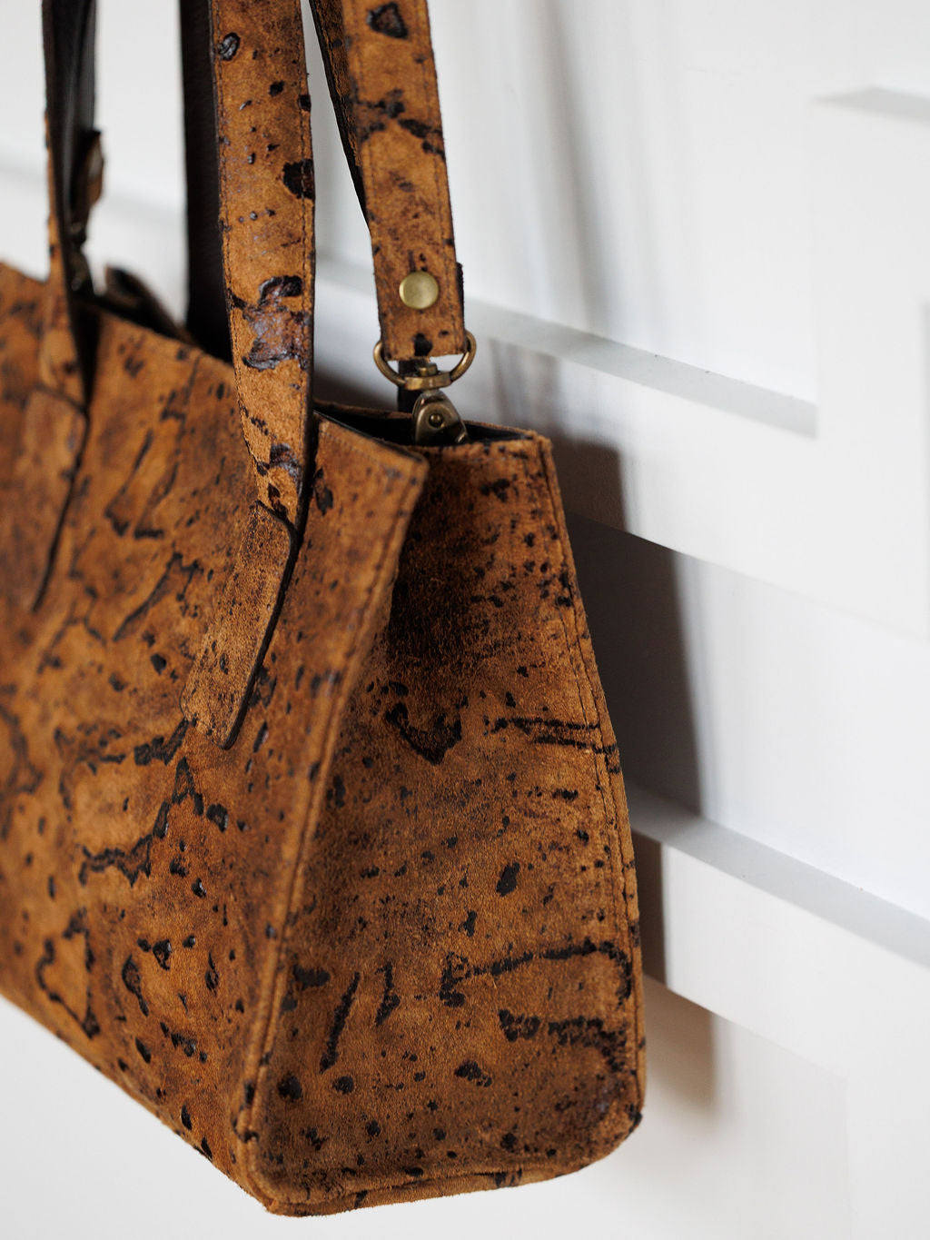 Close-up of The Nubuck by Savannah Sevens western life{&}style, a handmade brown cork and nubuck leather crossbody bag with dark natural patterns, sturdy handles, and a brass clasp, hanging on a white wall.