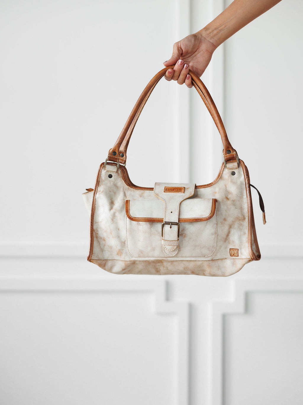 A hand holds The Rowley by Savannah Sevens western life{&}style, a white and brown marbled full grain leather handbag with tan handles, trim, front buckle pocket, and structured rectangular shape, set against a white paneled background.