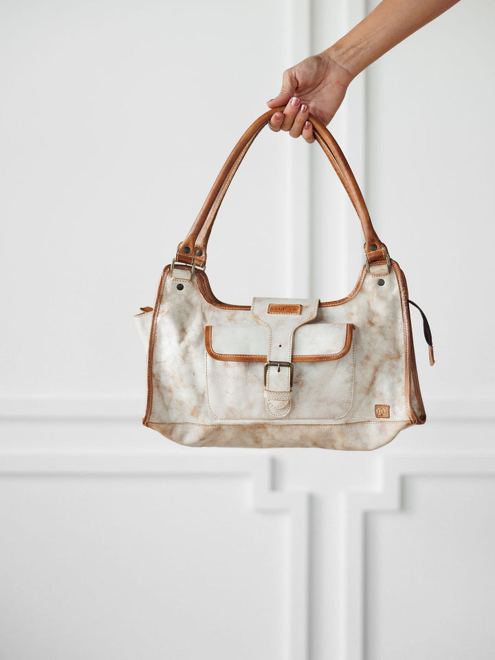 A hand holds The Rowley by Savannah Sevens western life{&}style, a white and brown marbled full grain leather handbag with tan handles, trim, front buckle pocket, and structured rectangular shape, set against a white paneled background.