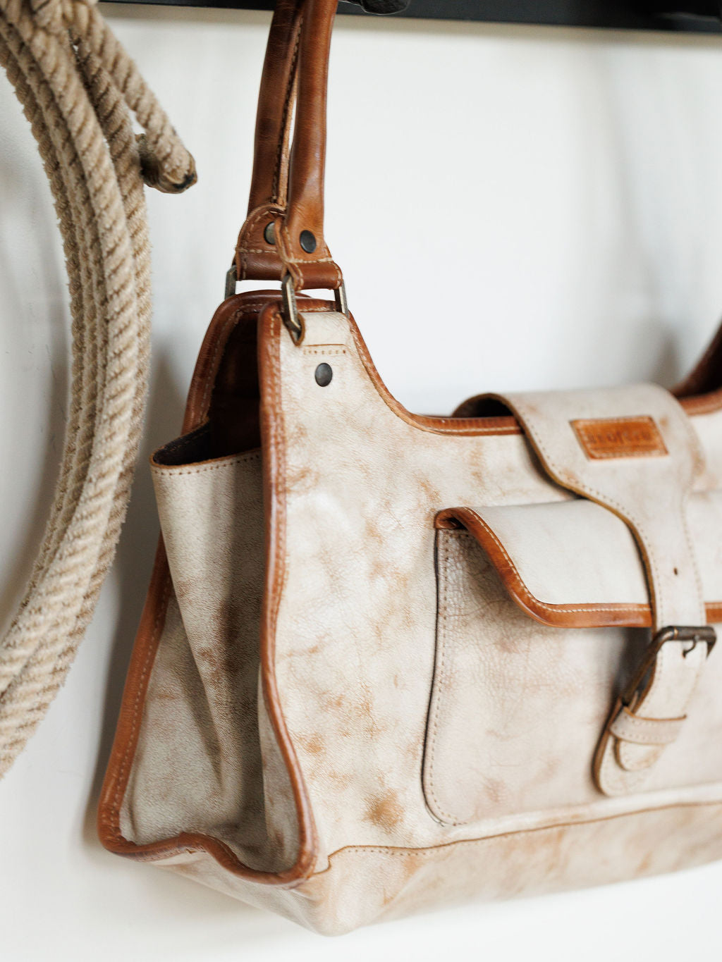 A close-up of The Rowley by Savannah Sevens western life{&}style, a rustic beige and brown full grain leather handbag with a front pocket and buckle, hanging by its handles next to a coiled rope on a white background.
