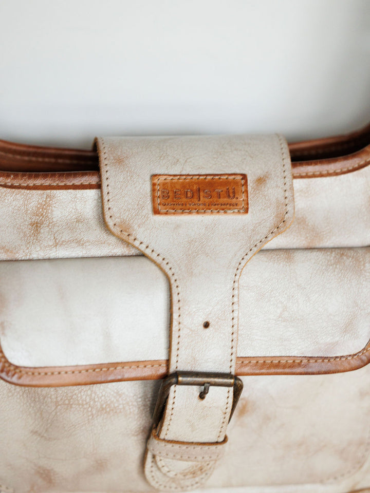 A close-up of The Rowley by Savannah Sevens western life{&}style, a rustic light beige leather handbag with brown trim and buckle. The flap features a rectangular patch, showcasing its full grain leather craftsmanship.