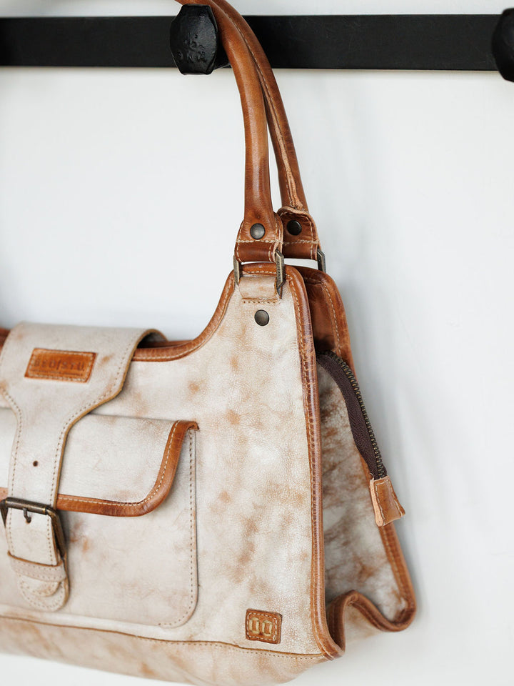 A close-up of The Rowley by Savannah Sevens western life{&}style, a rustic cream and brown full grain leather handbag featuring a front pocket and buckle detail, hanging from a black hook on a white wall.