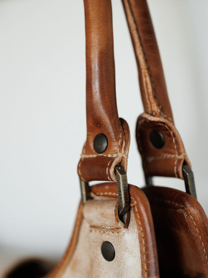 Close-up of two The Rowley bag handles by Savannah Sevens western life{&}style in brown full grain leather with dark metal hardware and rivets, showing textured leather and detailed stitching. Background is blurred.