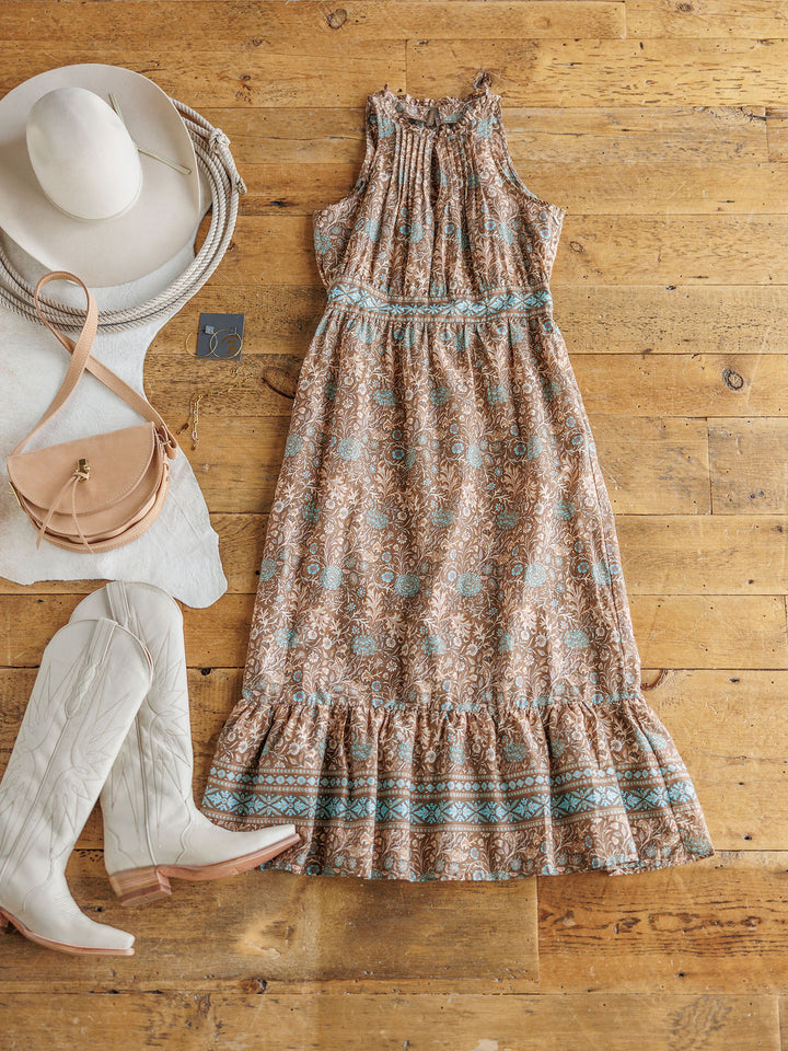 The Mayetta by Savannah Sevens western life{&}style, a western floral dress with blue accents, is styled with white cowboy boots, a tan crossbody bag, a wide-brimmed hat, and a silver necklace on a wooden floor.