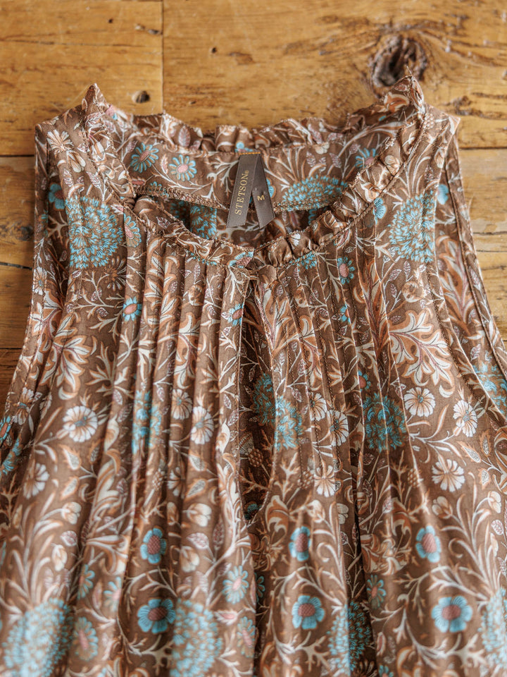 The Mayetta by Savannah Sevens western life{&}style is a sleeveless brown blouse with blue and white floral print, pleats, and a high neckline, displayed on rustic wood. A tag reading SEZANE is visible.