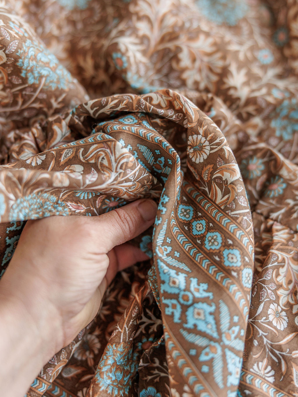 A hand gathers The Mayetta by Savannah Sevens western life{&}style, showcasing brown fabric detailed with intricate light blue and white western floral patterns.
