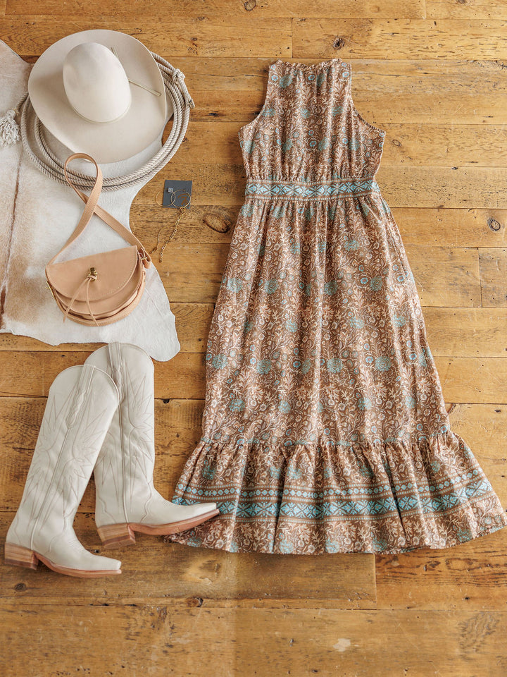 The Mayetta by Savannah Sevens western life{&}style, a sleeveless western floral dress with gathered waist and ruffled hem, is styled on a wooden floor with white cowboy boots, tan crossbody purse, white wide-brim hat, and coiled rope.
