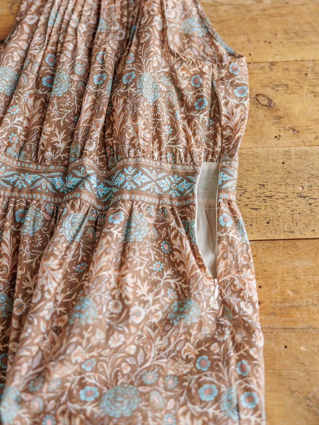 A close-up of The Mayetta dress by Savannah Sevens western life{&}style showcases blue and white floral designs, a geometric waistband, and a visible side pocket, all laid flat on a wooden surface.