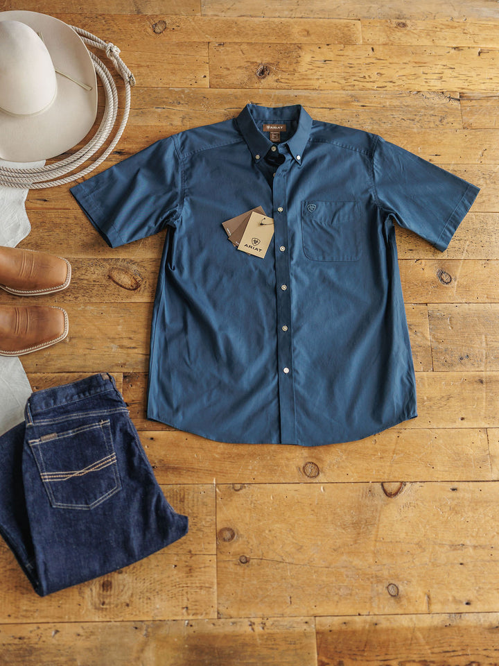 The Wellman men's short sleeve button-up shirt by Savannah Sevens western life&style in blue is 100% cotton with a classic fit, shown with tags on a wooden floor, alongside a white cowboy hat, brown boots, and blue jeans.