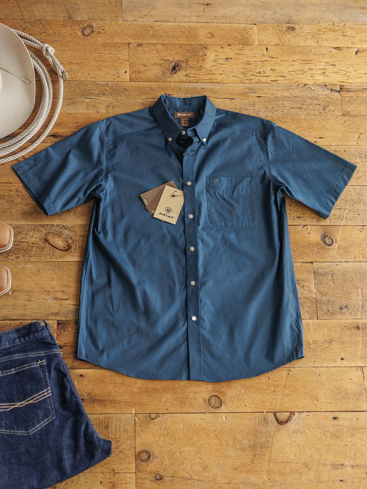 The Wellman Men's short sleeve 100% cotton blue button-up shirt by Savannah Sevens western life&style is laid flat with two tags near the front pocket, beside a white hat, denim jeans, and brown sandals on a wooden floor.