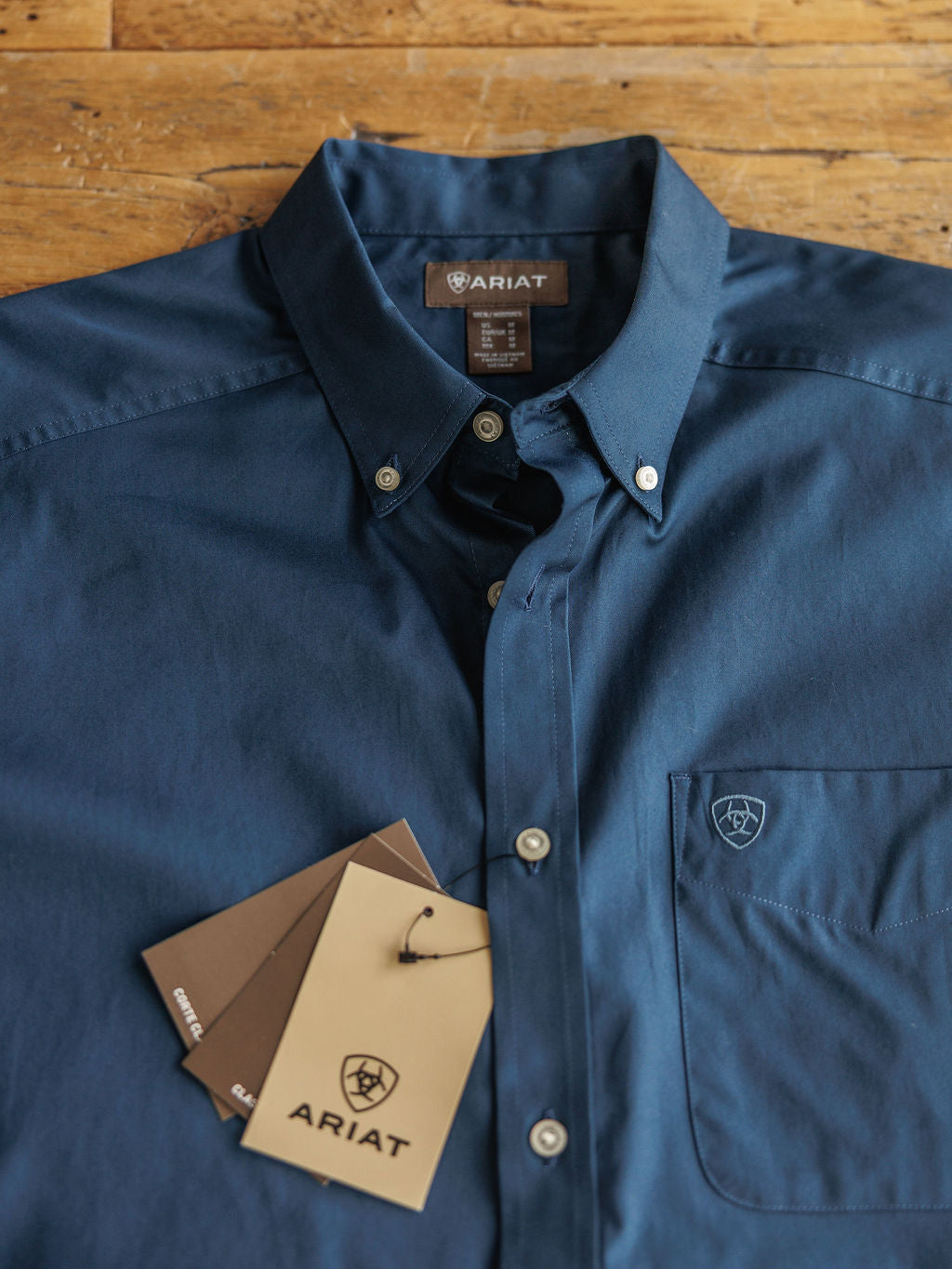 The Wellman men's short sleeve button-up shirt by Savannah Sevens western life&style in navy blue, 100% cotton with classic fit, front pocket, embroidered logo, and three branded tags attached near the pocket on a wooden surface.