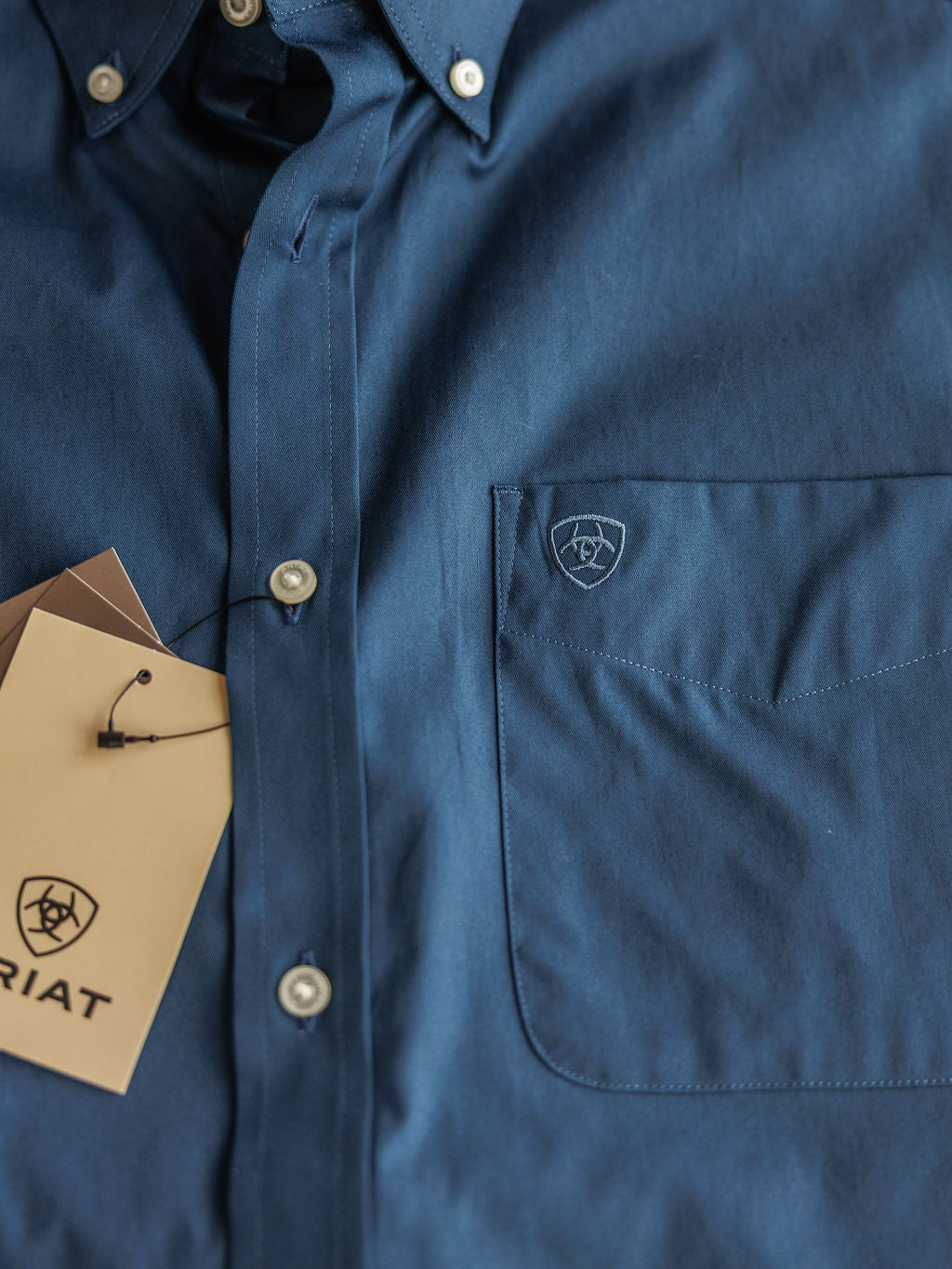 Close-up of The Wellman men's blue classic fit short sleeve button-up shirt by Savannah Sevens western life{&}style, featuring a front pocket, two tan tags by the buttons. Made from 100% cotton for comfort and style.