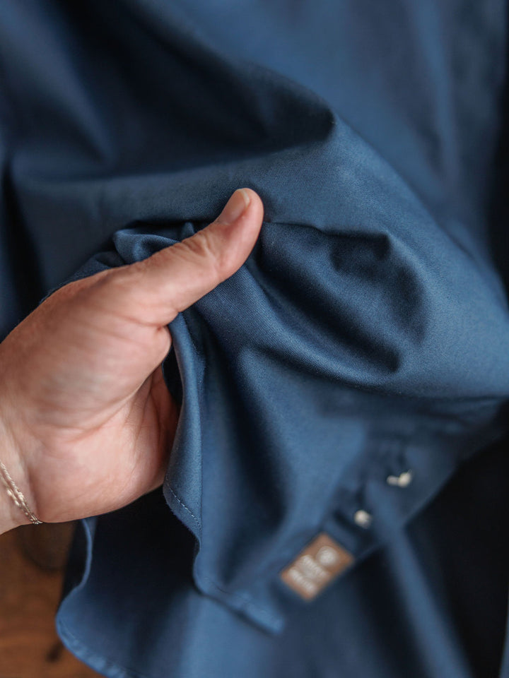 A hand gently scrunches smooth blue fabric from The Wellman {Men's} classic fit button-up by Savannah Sevens western life{&}style, highlighting its texture. A small brown tag with white text appears near the bottom.