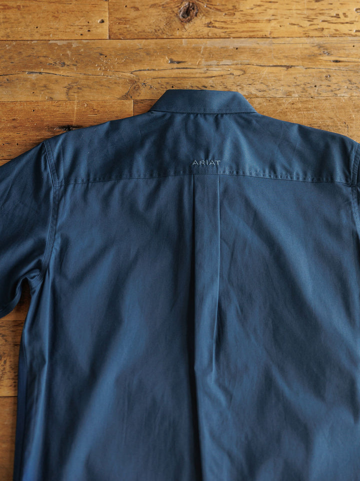 A navy blue classic fit button-up shirt, The Wellman {Men's} by Savannah Sevens western life{&}style, laid flat on a wooden surface with embroidered detail on the upper back.