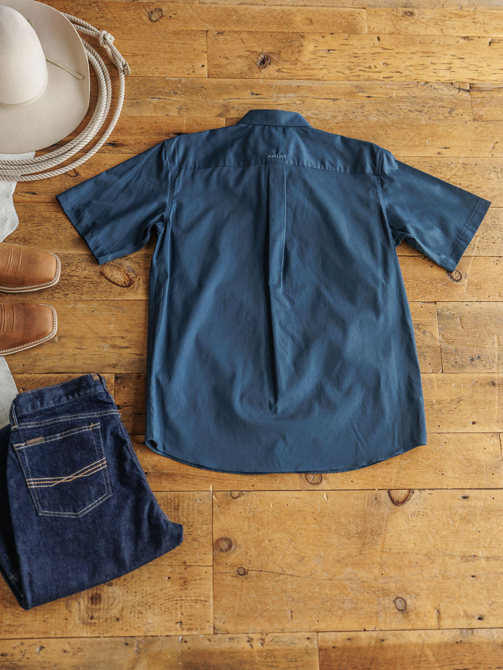 The Wellman men's short sleeve button up shirt by Savannah Sevens western life{&}style is laid flat with a beige cowboy hat, brown boots, and dark blue jeans for a timeless, classic Western look.