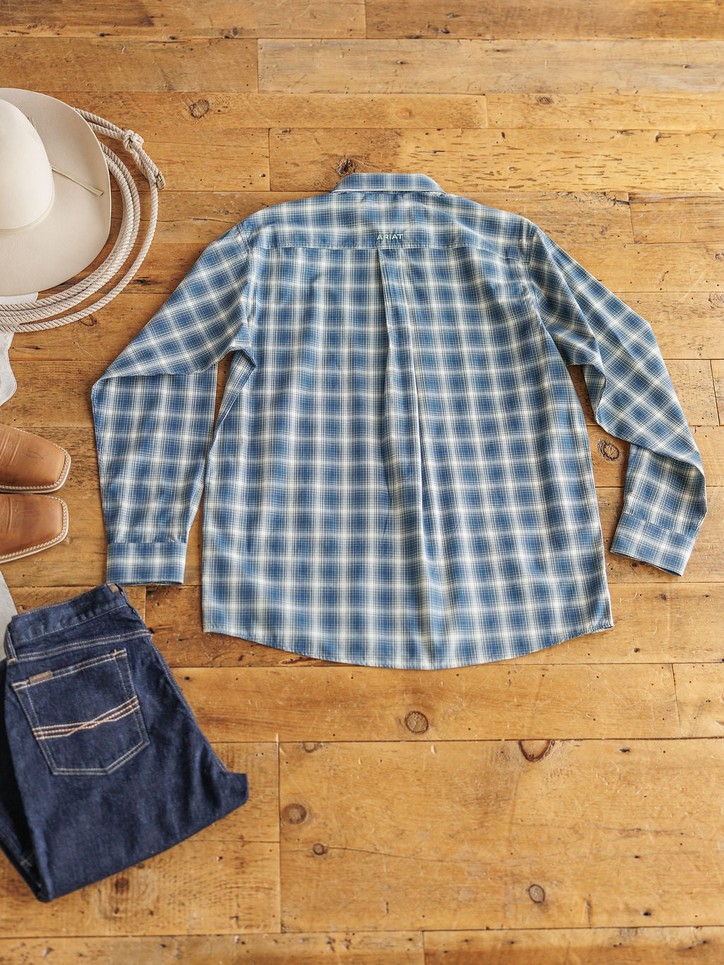 The Davis {Men’s} plaid long sleeve shirt by Savannah Sevens western life{&}style, featuring sun protection, is laid flat and back-facing on a wooden floor with folded blue jeans, brown boots, a coiled rope, and a white cowboy hat.