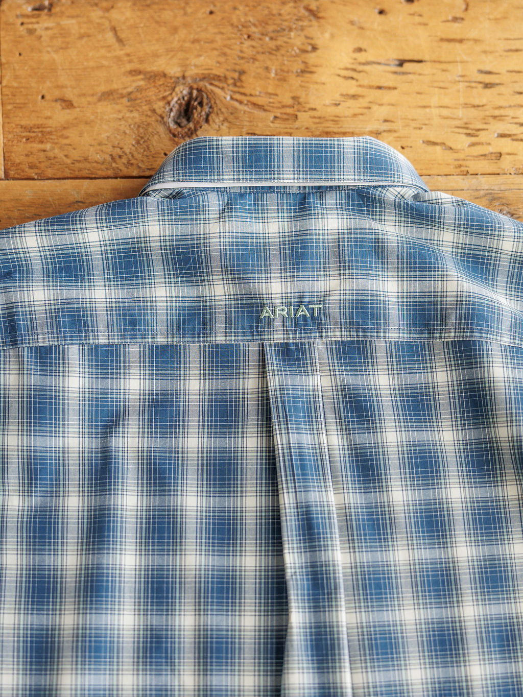 The Davis men's blue plaid long sleeve shirt by Savannah Sevens western life{&}style features white and navy stripes, shown from the back with yellow embroidery. Moisture wicking; laid flat on a wooden surface.