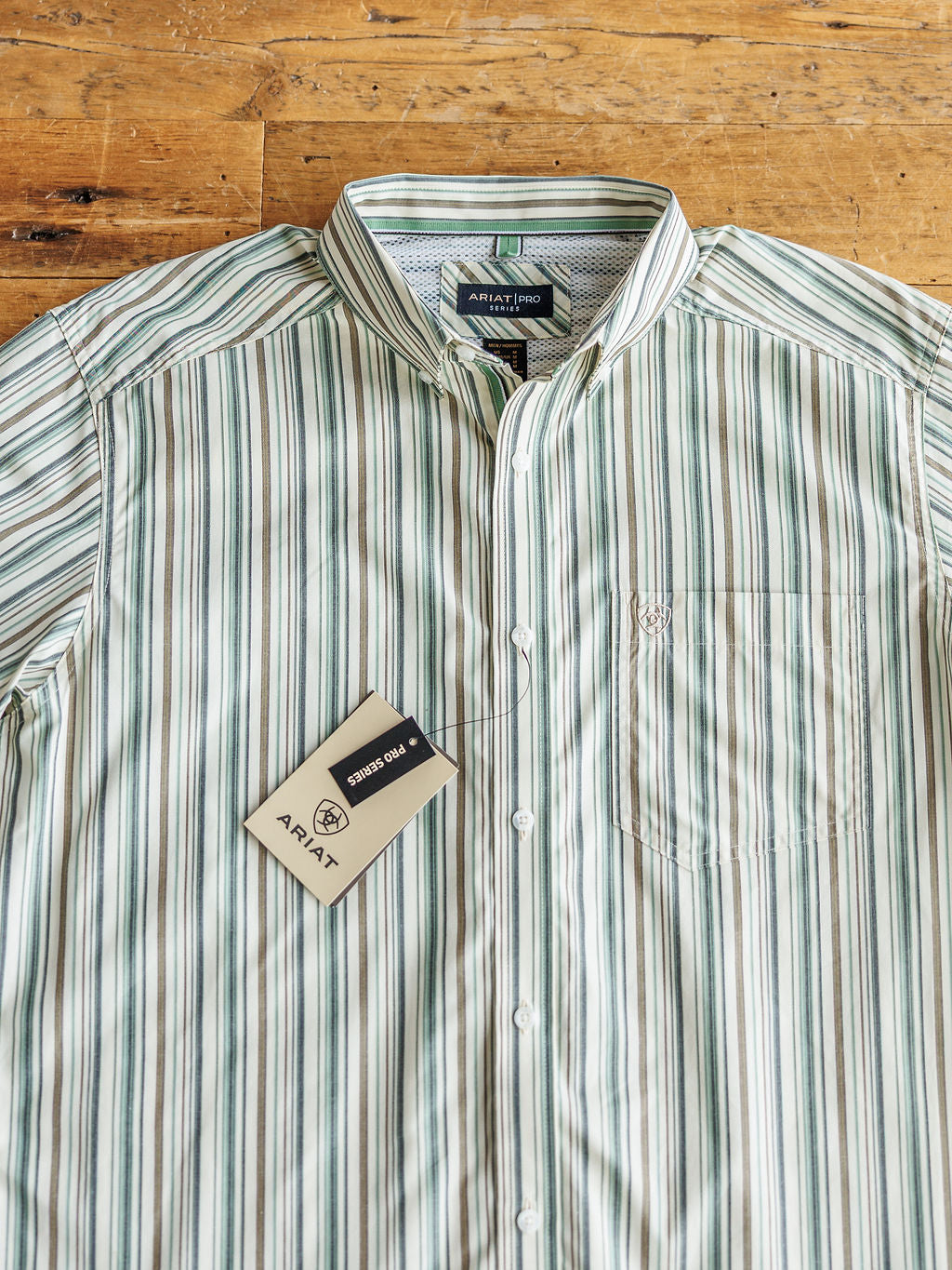 A green and white striped The Austwell men's shirt by Savannah Sevens western life&style is laid flat on a wooden surface. This short sleeve, collared button-up features a chest pocket and a branded tag.
