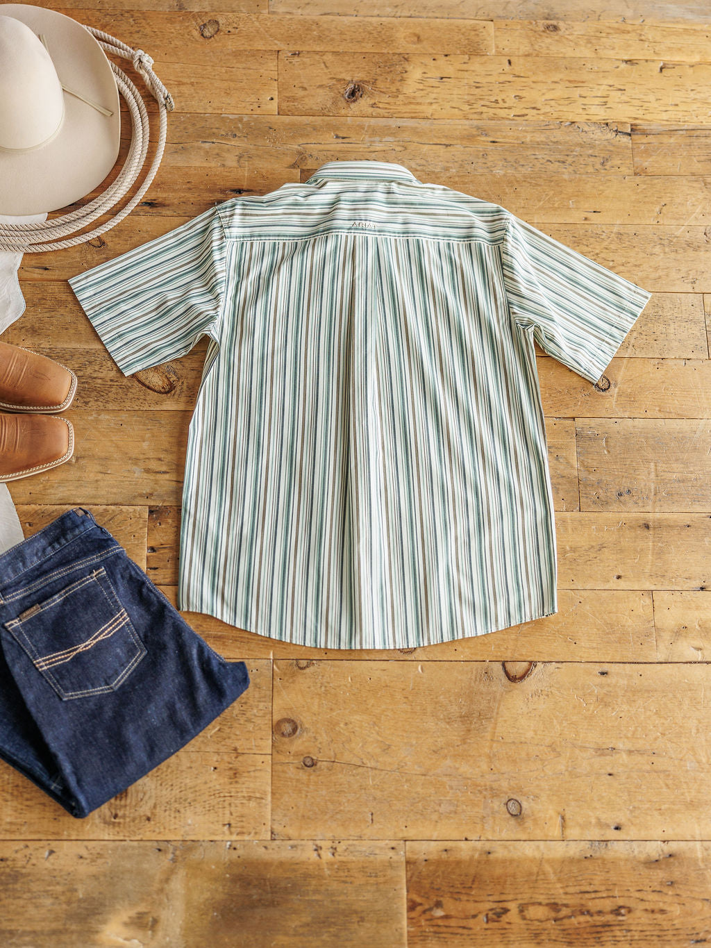 The Austwell Men's shirt by Savannah Sevens western life&style in light green and white stripes is laid flat on a wooden floor, paired with a tan cowboy hat, brown boots, and folded dark blue jeans.