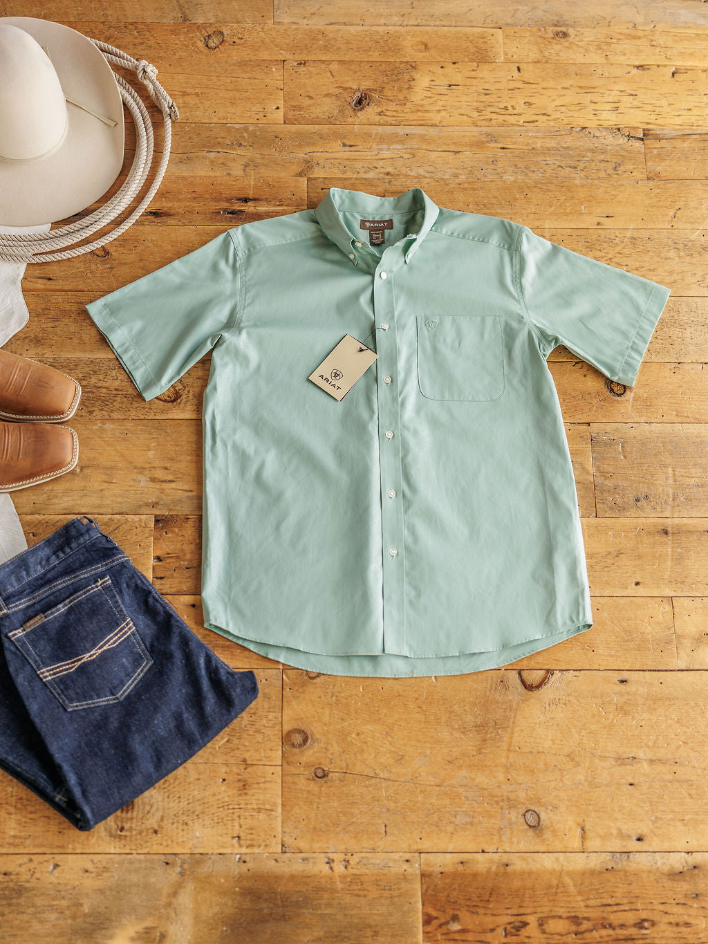 The Williamsburg men's sage short sleeve button up by Savannah Sevens western life{&}style is laid flat with a white cowboy hat, tan shoes, blue jeans, and features a front pocket and attached tag. Classic fit cotton shirt displayed on wooden floor.