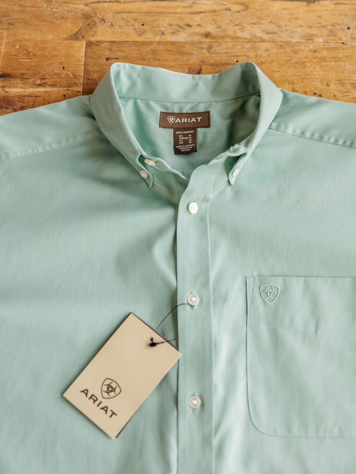 A sage green men’s classic fit cotton shirt, The Williamsburg by Savannah Sevens western life&style, is neatly laid out on a wooden surface with the brand tag attached to the chest pocket and label visible inside the collar.