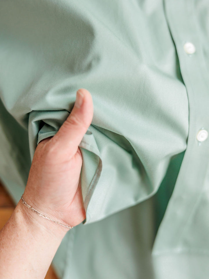 A hand gently holds the soft fabric of The Williamsburg {Men's} sage short sleeve button-up by Savannah Sevens western life{&}style, showing its texture and drape. White buttons and a silver bracelet are visible.