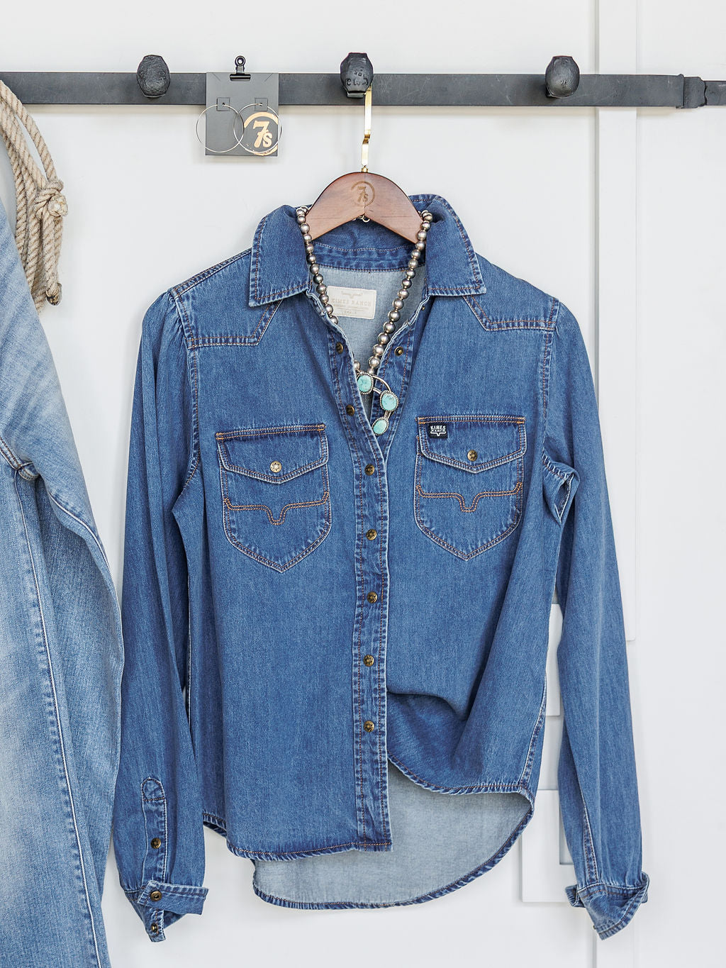 The Dalles by Savannah Sevens western life{&}style, a women's relaxed fit denim snap button top with front pockets, hangs on a hook against a white wall. A beaded necklace is on the hanger; part of a denim jacket appears at left.