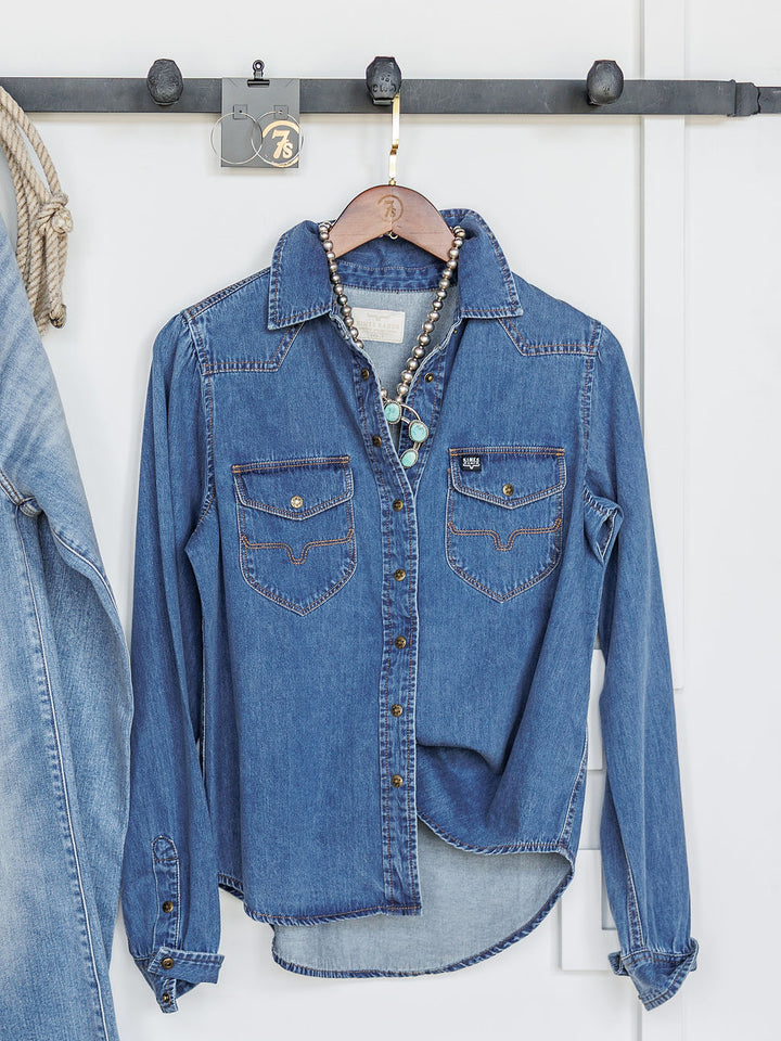 The Dalles by Savannah Sevens western life{&}style, a women's relaxed fit denim snap button top with front pockets, hangs on a hook against a white wall. A beaded necklace is on the hanger; part of a denim jacket appears at left.