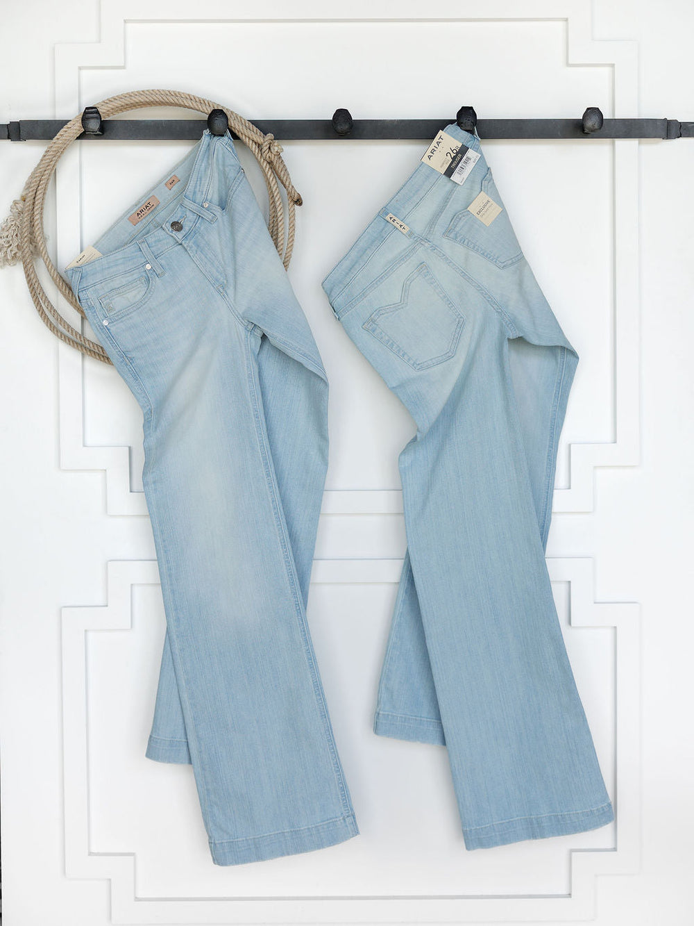 Two pairs of The Dani Perfect Rise Trouser Jean *Exclusive* by Savannah Sevens western life{&}style hang on a black rod against a white wall, with a coiled rope also hanging on the left side.