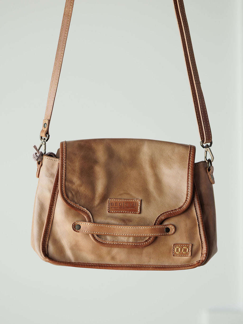 The Lomita by Savannah Sevens western life{&}style, a rustic oat leather crossbody with flap closure, decorative stitching, and a long adjustable strap, hangs against a plain light background.