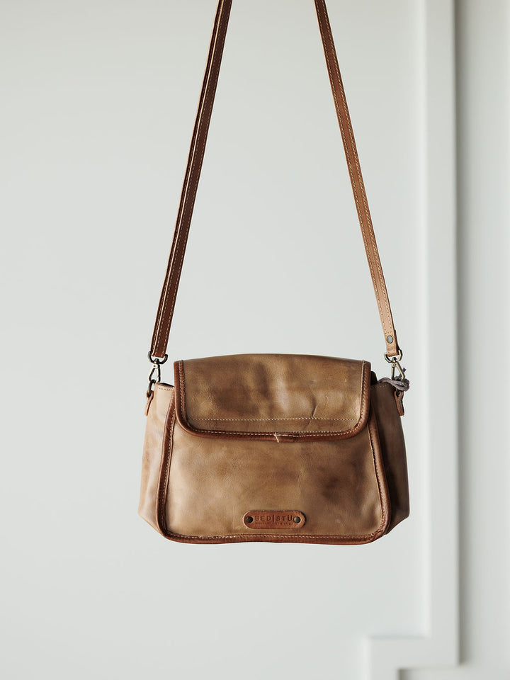 The Lomita crossbody bag by Savannah Sevens western life{&}style, crafted in rustic oat leather with a long adjustable strap, hangs against a neutral background and features a flap closure and a small patch label on the front.