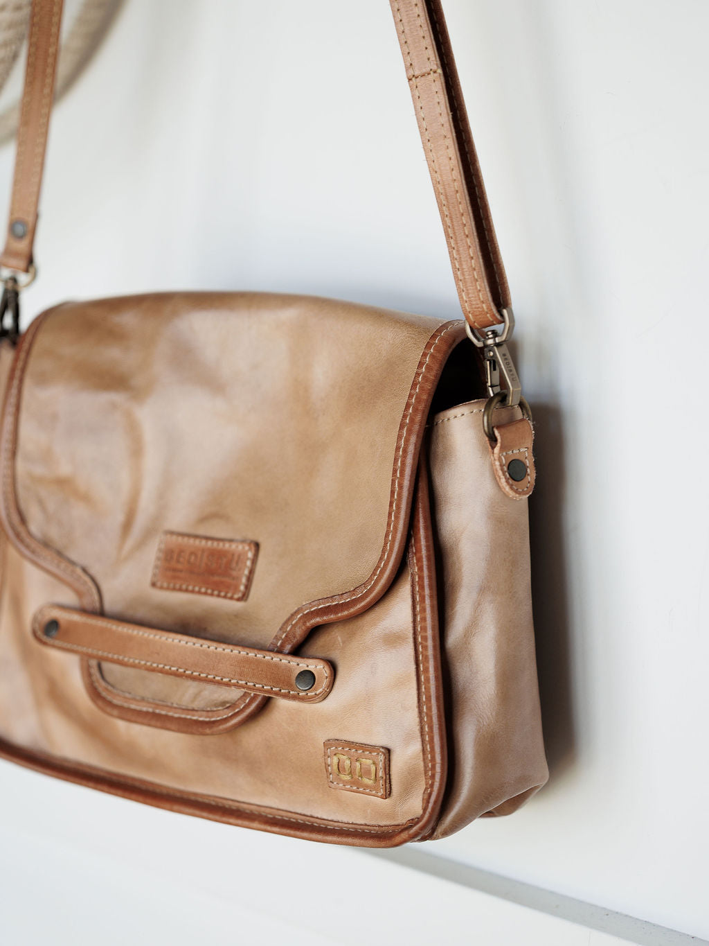 The Lomita by Savannah Sevens western life{&}style is a leather crossbody bag in rustic oat with brown trim and stitching, shown against a white background. It features a front flap with rivet accents and a stitched patch on the lower right corner.