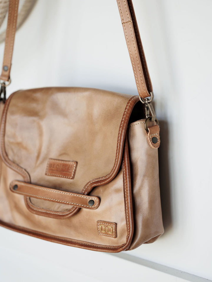 The Lomita by Savannah Sevens western life{&}style is a leather crossbody bag in rustic oat with brown trim and stitching, shown against a white background. It features a front flap with rivet accents and a stitched patch on the lower right corner.