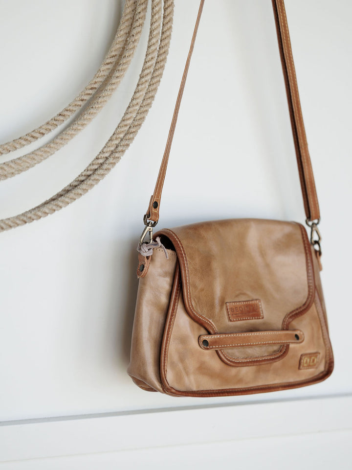 The Lomita leather crossbody bag by Savannah Sevens western life{&}style in rustic oat, featuring a buckle and stitched details, hangs on a wall beside a coiled beige rope.