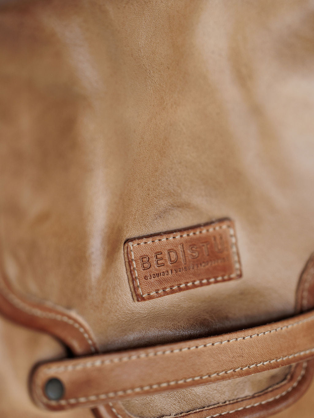 Close-up of The Lomita crossbody by Savannah Sevens western life{&}style in rustic oat leather, showcasing a stitched leather label with the brand name, visible quality texture, a leather strap, and metal rivet.