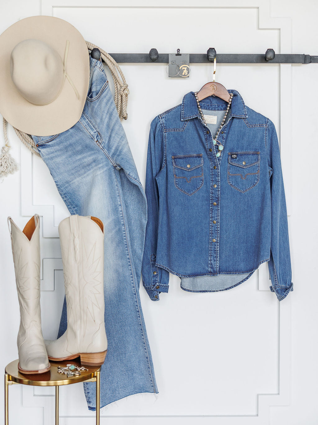 The Dalles by Savannah Sevens western life{&}style—a beige cowboy hat, women's relaxed fit denim jeans, and a western yoke denim shirt—hang on a black rack as white cowboy boots rest on a gold table against a clean white wall.