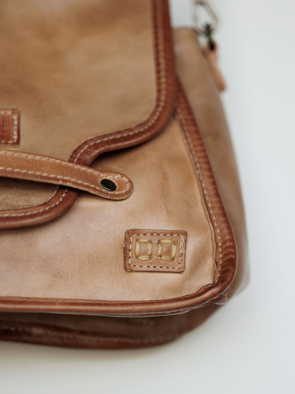 Close-up of The Lomita by Savannah Sevens western life{&}style, a leather crossbody bag in rustic oat with visible stitching and a metal snap button on the flap, resting on a white surface.